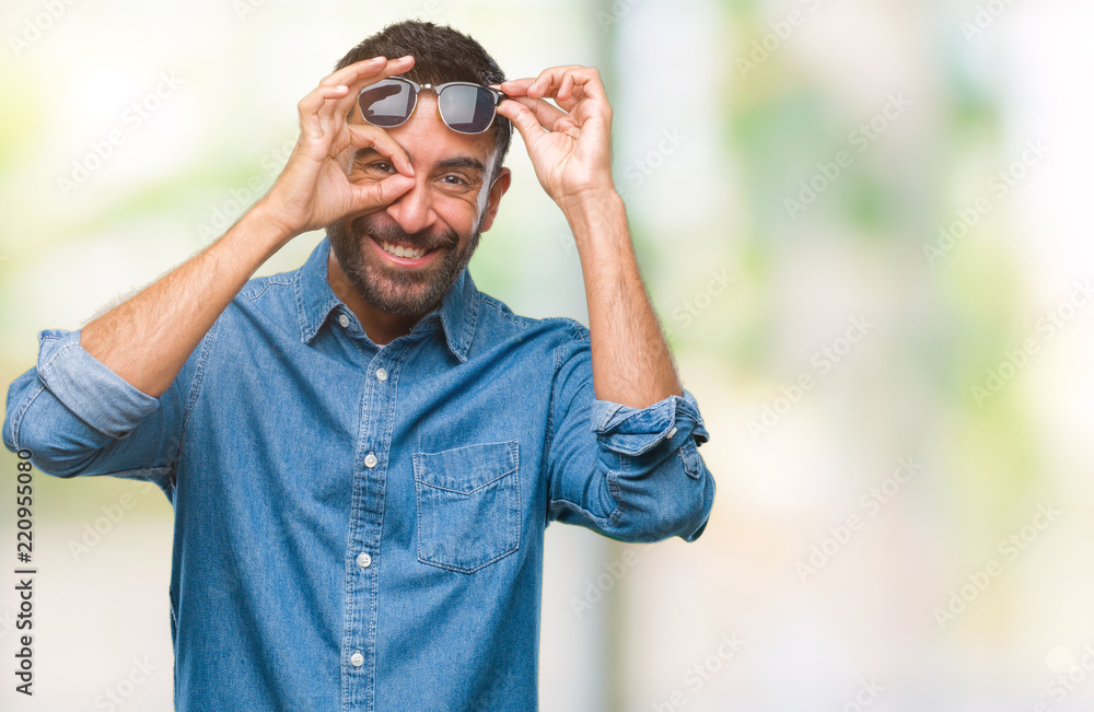 Naklejka premium Adult hispanic man wearing sunglasses over isolated background doing ok gesture with hand smiling, eye looking through fingers with happy face.