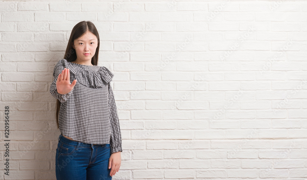 Young Chinese woman over brick wall with open hand doing stop sign with serious and confident expression, defense gesture Young Chinese woman over brick wall with open hand doing stop sign with serious and confident expression, defense gesture
