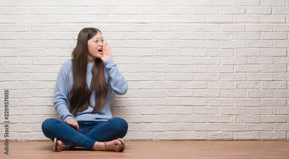 Young Chinese woman sitting on the floor over brick wall shouting and screaming loud to side with hand on mouth. Communication concept. Young Chinese woman sitting on the floor over brick wall shouting and screaming loud to side with hand on mouth. Communication concept.