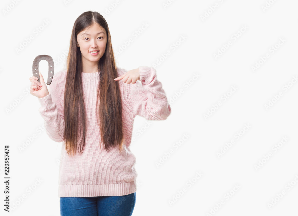 Young Chinese woman over isolated background holding horseshoe talisman with surprise face pointing finger to himself Young Chinese woman over isolated background holding horseshoe talisman with surprise face pointing finger to himself