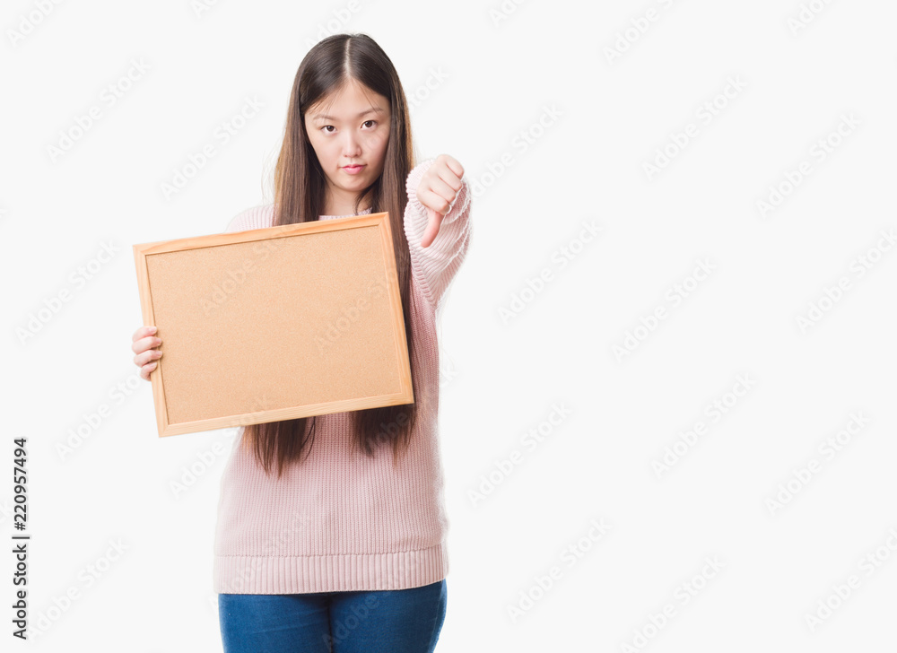 Young Chinese woman over isolated background holding corkboard with angry face, negative sign showing dislike with thumbs down, rejection concept Young Chinese woman over isolated background holding corkboard with angry face, negative sign showing dislike with thumbs down, rejection concept