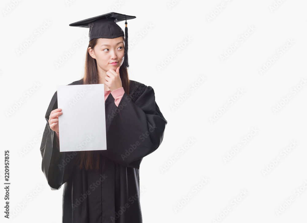 Young Chinese woman wearing graduate uniform holding paper degree serious face thinking about question, very confused idea Young Chinese woman wearing graduate uniform holding paper degree serious face thinking about question, very confused idea