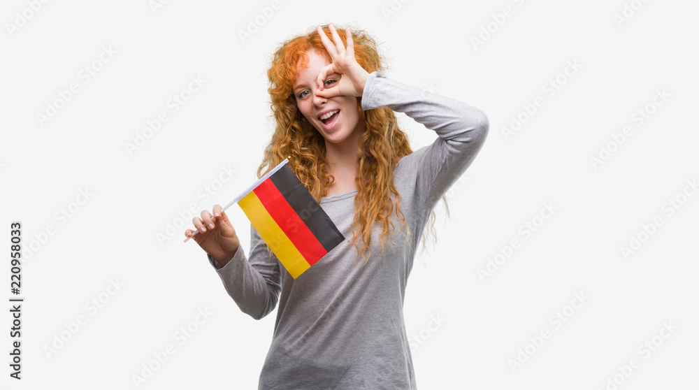 Young redhead woman holding flag of Germany with happy face smiling doing ok sign with hand on eye looking through fingers Young redhead woman holding flag of Germany with happy face smiling doing ok sign with hand on eye looking through fingers
