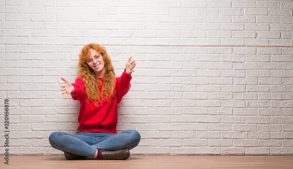 Young redhead woman sitting over brick wall looking at the camera smiling with open arms for hug. Cheerful expression embracing happiness. Young redhead woman sitting over brick wall looking at the camera smiling with open arms for hug. Cheerful expression embracing happiness.