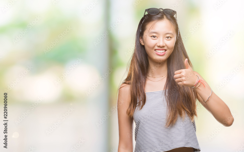 Young asian woman wearing sunglasses over isolated background doing happy thumbs up gesture with hand. Approving expression looking at the camera with showing success.