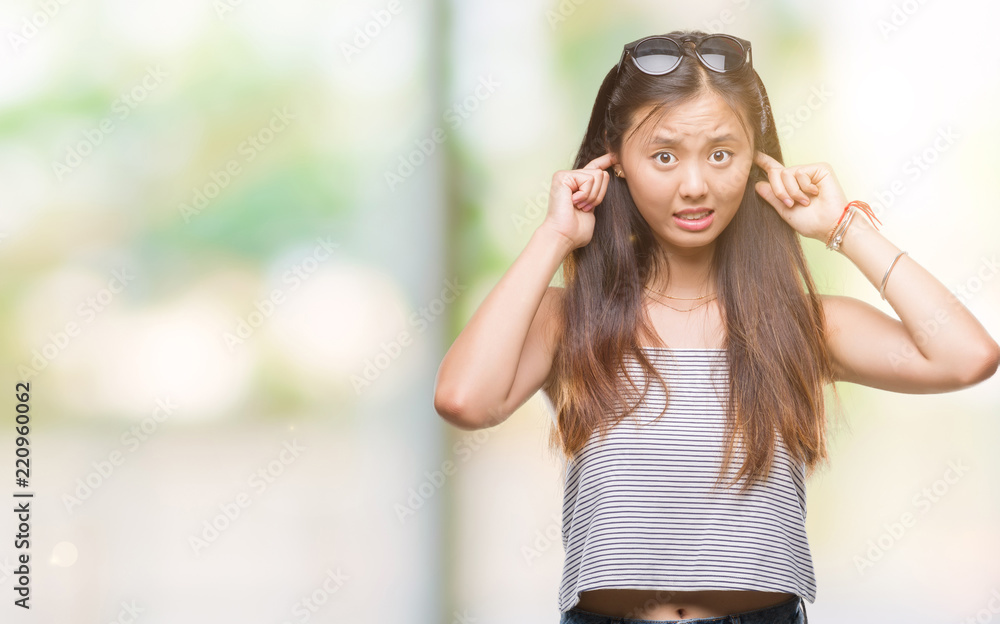 Young asian woman wearing sunglasses over isolated background covering ears with fingers with annoyed expression for the noise of loud music. Deaf concept.