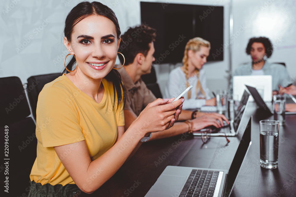 © LIGHTFIELD STUDIOS - smiling young businesswoman with smartphone and her partners having discussion behind at modern office