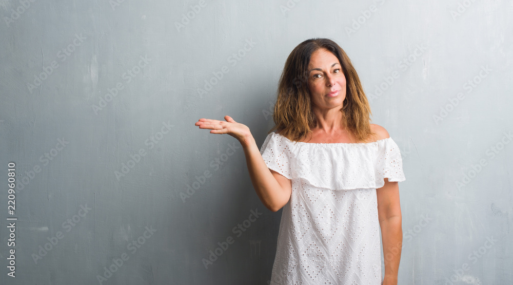 Middle age hispanic woman standing over grey grunge wall smiling cheerful presenting and pointing with palm of hand looking at the camera. Middle age hispanic woman standing over grey grunge wall smiling cheerful presenting and pointing with palm of hand looking at the camera.
