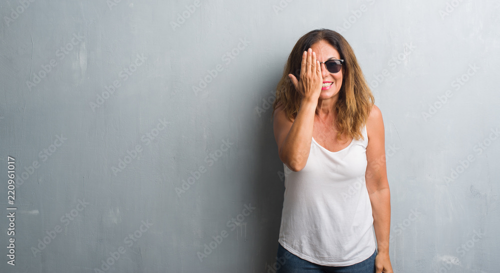 Middle age hispanic woman over grey wall wearing sunglasses covering one eye with hand with confident smile on face and surprise emotion.