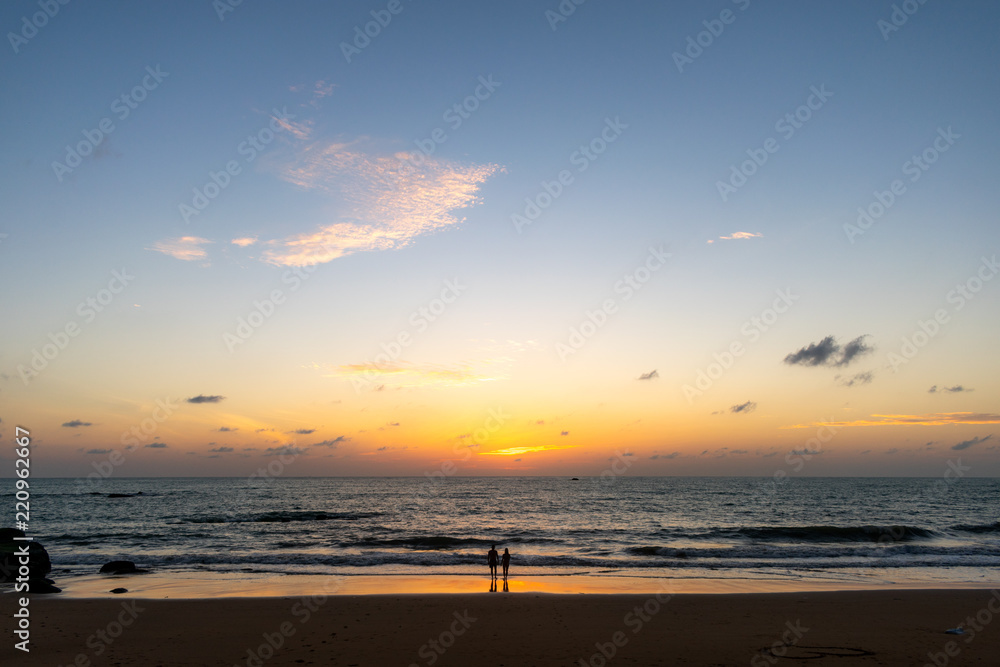 A landscape of a beach just after sunset with two people silhouette standing on the sand by, water in the background