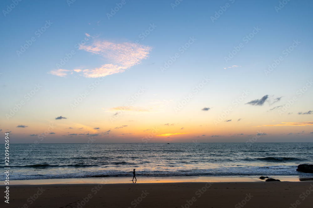 A landscape of a beach just after sunset with a person silhouette walking by, water in the background