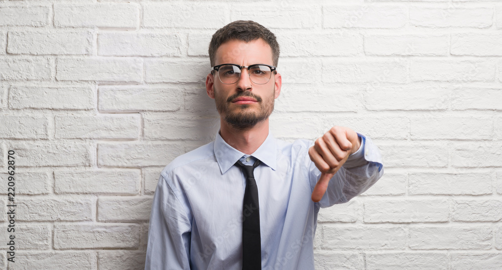Young adult business man standing over white brick wall with angry face, negative sign showing dislike with thumbs down, rejection concept Young adult business man standing over white brick wall with angry face, negative sign showing dislike with thumbs down, rejection concept