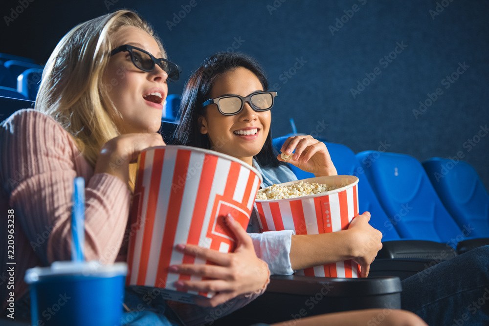 Naklejka premium emotional multiracial women in 3d glasses with popcorn watching film together in movie theater