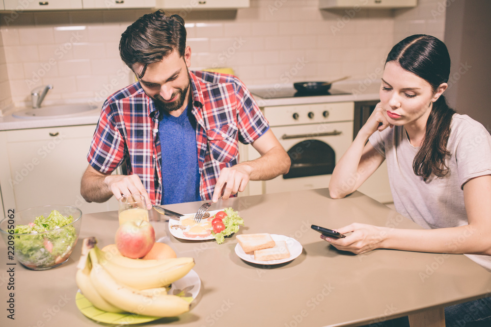 Happy guy sits and table and eats omlet. He cuts food into pieces with knife and fork. Girl looks at phone she holds in hands. She is bored and tired.