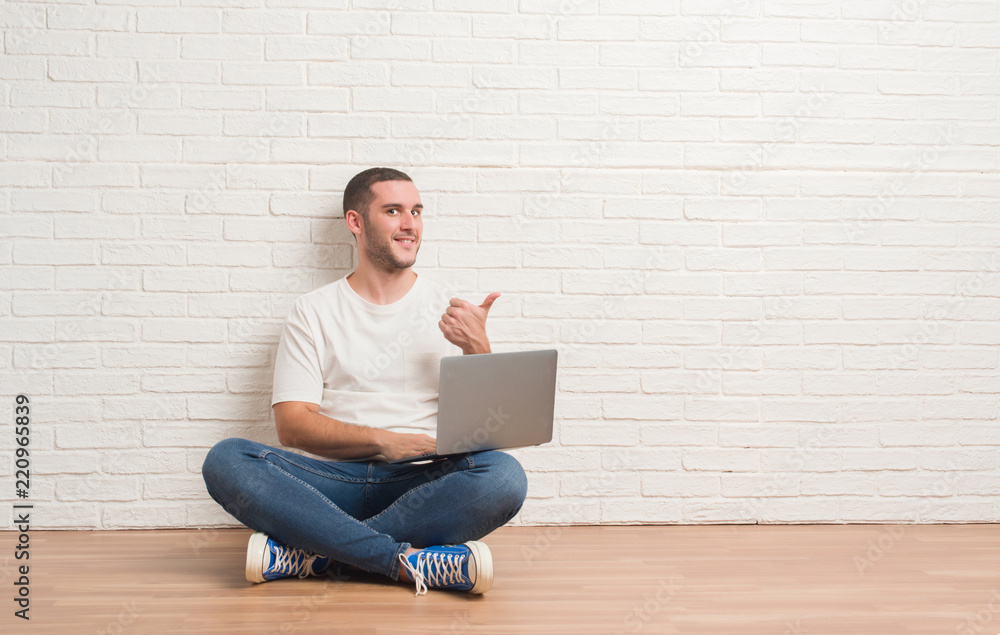 Young caucasian man sitting over white brick wall using computer laptop pointing and showing with thumb up to the side with happy face smiling