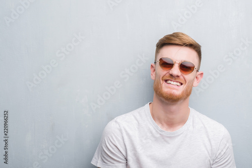 Young redhead man over grey grunge wall wearing retro sunglasses with a happy face standing and smiling with a confident smile showing teeth