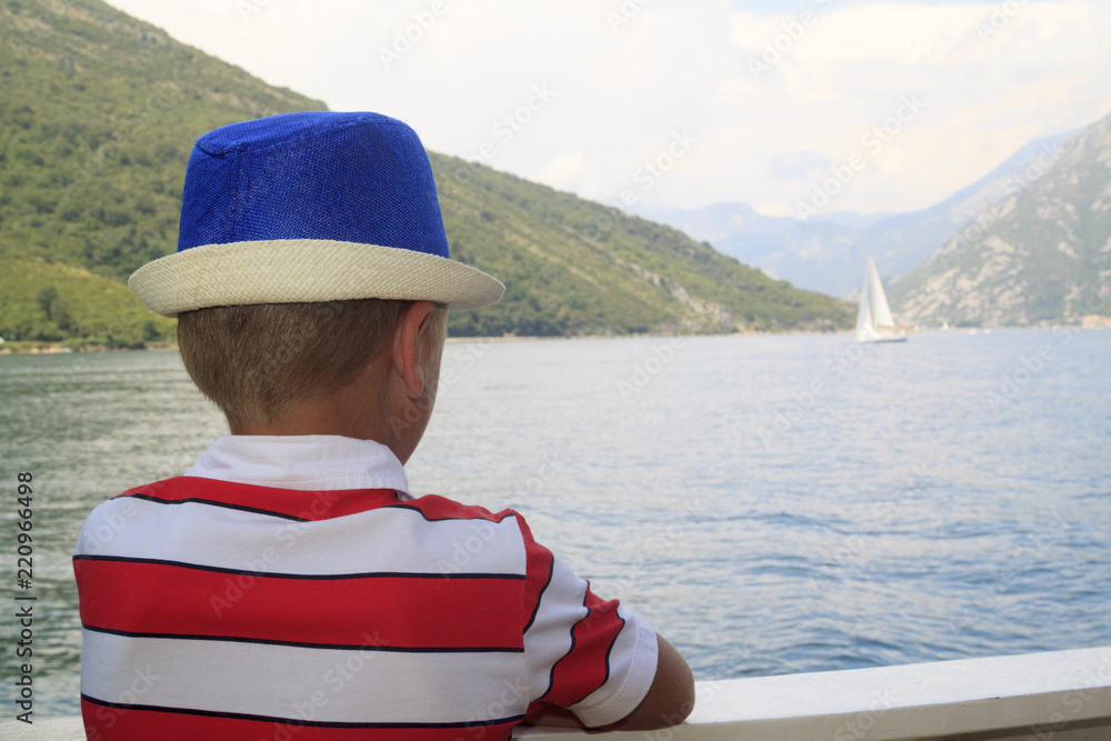 boy in a blue hat sails on a ferry across the Bay of Kotor and enthusiastically looks into the distance to the mountains and a sailing boat