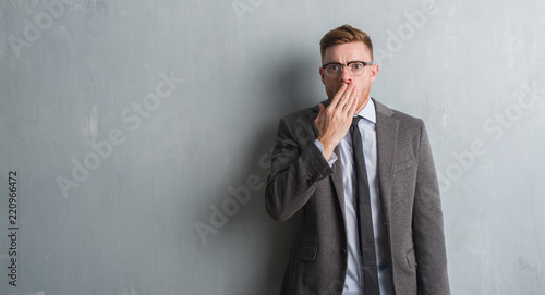 Young redhead elegant business man over grey grunge wall cover mouth with hand shocked with shame for mistake, expression of fear, scared in silence, secret concept