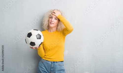 Young blonde woman over grunge grey background holding soccer football ball stressed with hand on head, shocked with shame and surprise face, angry and frustrated. Fear and upset for mistake.