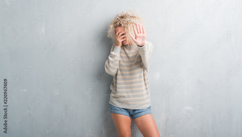 Young blonde woman with curly hair over grunge grey background covering eyes with hands and doing stop gesture with sad and fear expression. Embarrassed and negative concept. Young blonde woman with curly hair over grunge grey background covering eyes with hands and doing stop gesture with sad and fear expression. Embarrassed and negative concept.