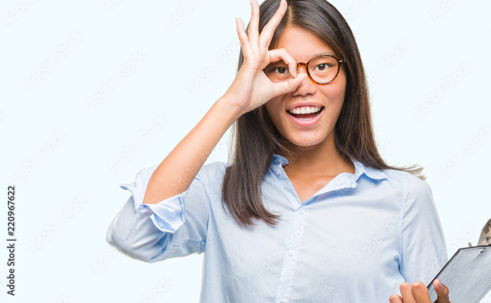 Young asian business woman over isolated background holding clipboard with happy face smiling doing ok sign with hand on eye looking through fingers