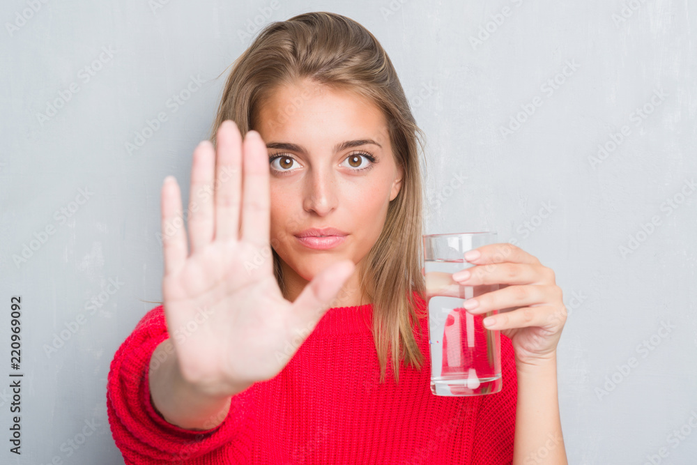 Beautiful young woman over grunge grey wall drinking a glass of water with open hand doing stop sign with serious and confident expression, defense gesture Beautiful young woman over grunge grey wall drinking a glass of water with open hand doing stop sign with serious and confident expression, defense gesture
