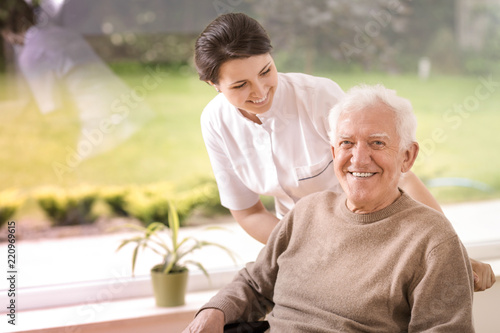 Friendly nurse supporting disabled smiling elderly man in the nursing house