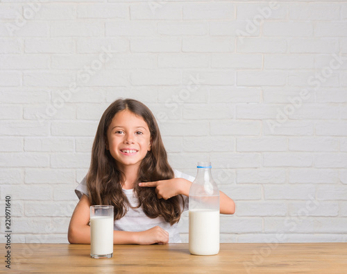 Young hispanic kid sitting on the table drinking a glass of milk with surprise face pointing finger to himself