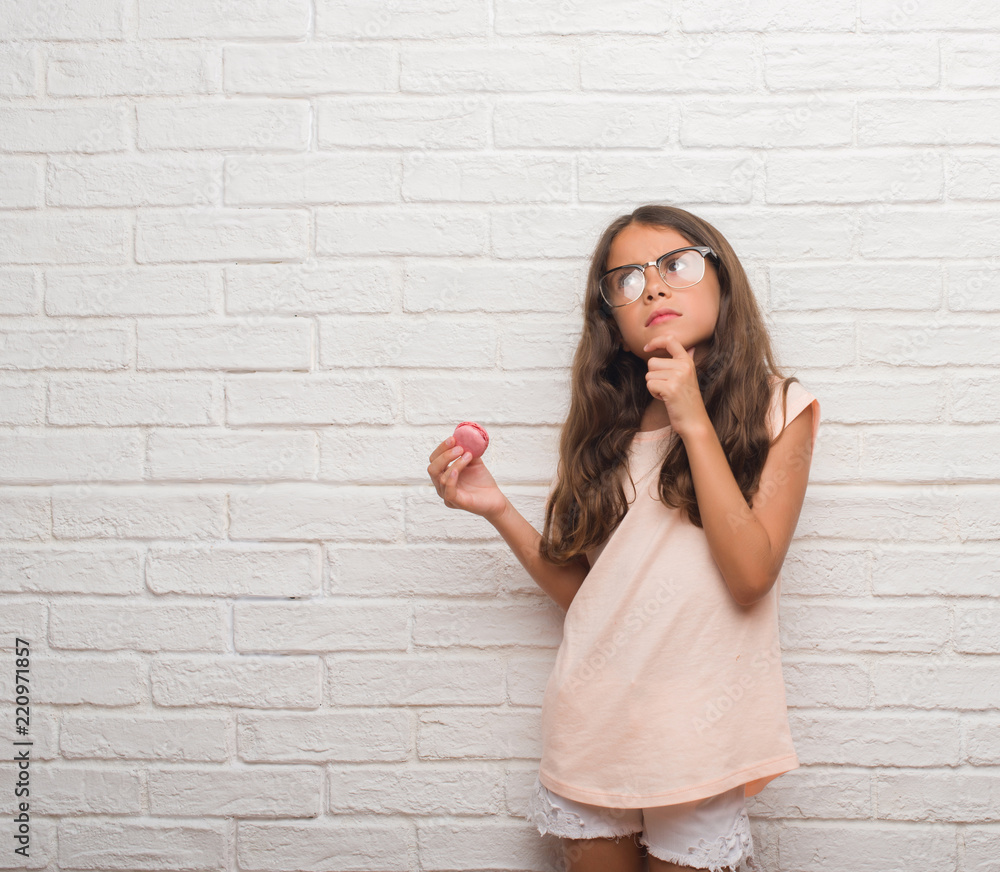 Young hispanic kid over white brick wall eating pink macaron serious face thinking about question, very confused idea Young hispanic kid over white brick wall eating pink macaron serious face thinking about question, very confused idea