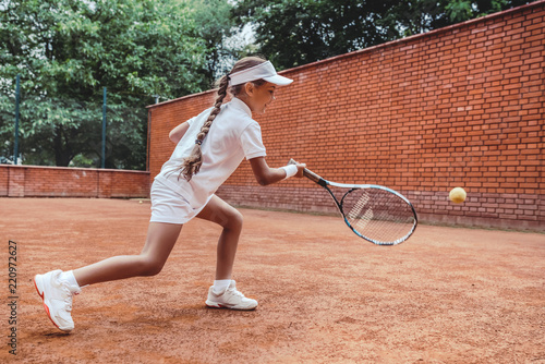 Child playing tennis on outdoor clay court. Full length shot of a tanned little girl on tennis court. Girl child playing tennis against the wall. Summer activities for children. © HBS