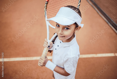 Tennis is my favorite game! Portrait of a pretty sporty child with a tennis racket. Little girl smiling at camera on tennis court. Training for young kid. © HBS