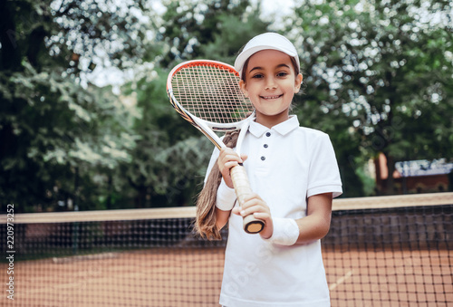 I choose healthy way with tennis! Portrait of pretty sporty child with tennis racquet. Cute little girl athlete on tennis court. Tennis player kid in white sportswear © HBS