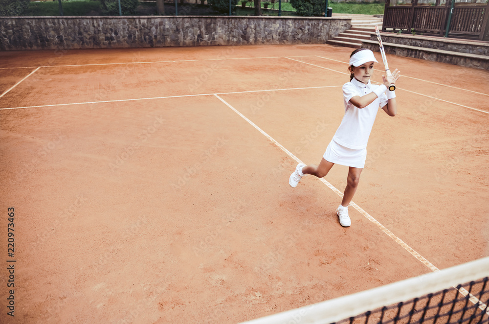 Obraz premium Child learning to play tennis in the sport club. Full length shot of a little girl tennis player on clay court. Sporty child girl hits the ball with a racket. Tennis training for young kid.