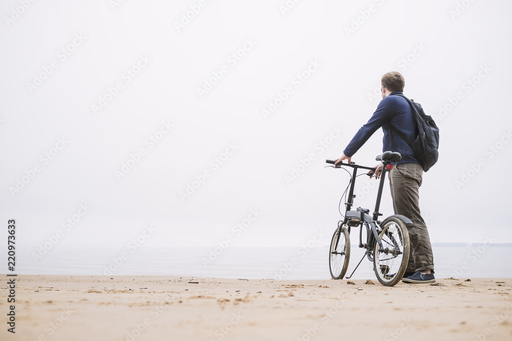 Fototapeta premium A young man with a backpack standing with the bike on the beach looking at the sea. 