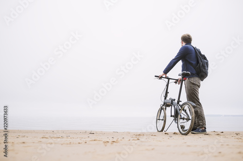 A young man with a backpack standing with the bike on the beach looking at the sea.
