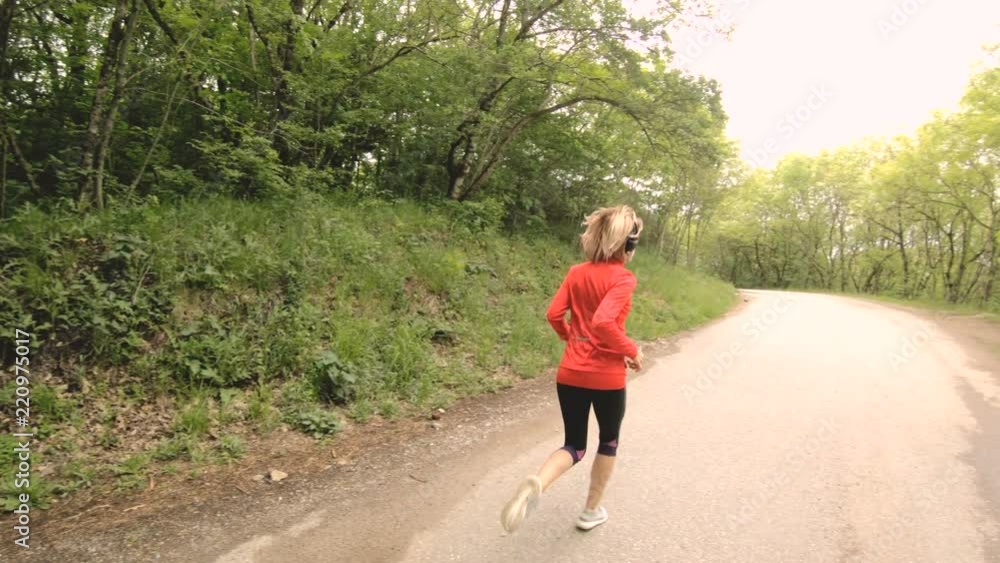 Young attractive woman in headphones changing the settings on a smart watch in front of or psole jogging on the road in a green forest Young attractive woman in headphones changing the settings on a smart watch in front of or psole jogging on the road in a green forest