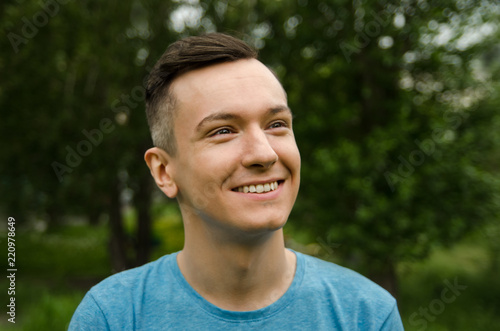 Young smiling guy looks aside on a green grass and trees background.