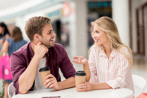 happy woman with paper cup of coffee gesturing by hand and talking to boyfriend at table in cafe