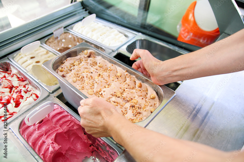 Confectioner is working at ice cream factory is holding, carrying creamy ice cream of hazelnut flavors with pieces, chunks and fruit of hazelnut in serving steel container, she is putting in fridge.