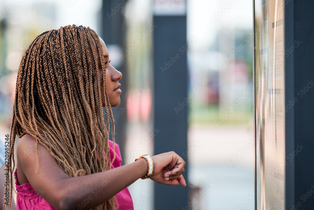 Young African woman checking plan and waiting at city bus stop