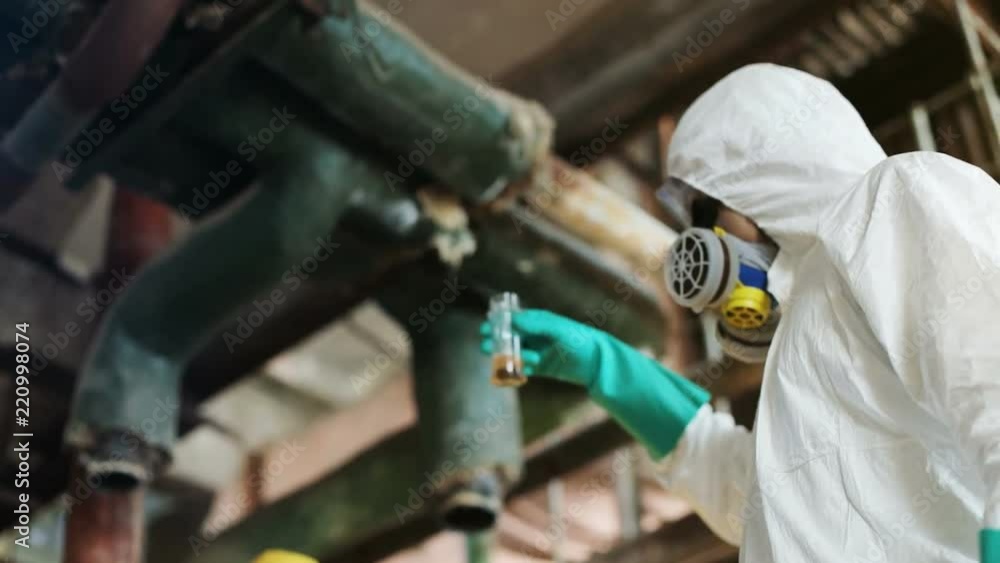 Caucasian man in white hazmat suit and respirator mask holding glassware in hand, shaking liquid. Two scientists at work. Indoors. Caucasian man in white hazmat suit and respirator mask holding glassware in hand, shaking liquid. Two scientists at work. Indoors.