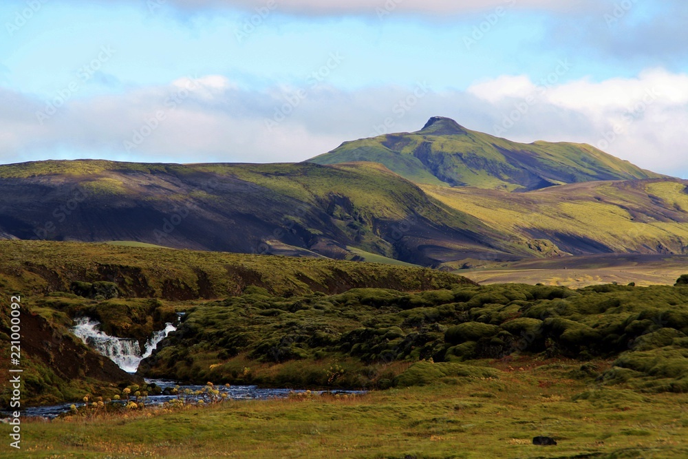 Paisaje de montañas de origen volcánico verdes, sin árboles y con cascada, en Islandia.