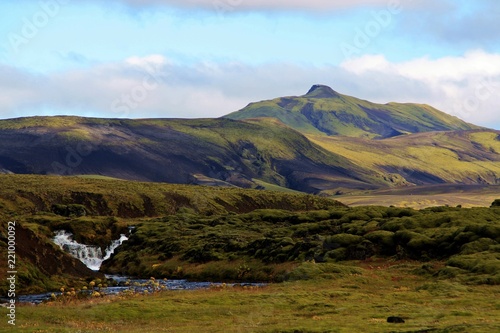 Paisaje de montañas de origen volcánico verdes, sin árboles y con cascada, en Islandia.
