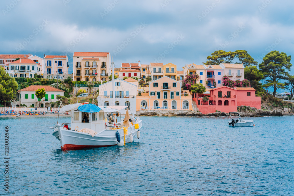 Fishing boat at anchor in blue sea bay of Assos village. Vivid colored houses with clouds in background, Kefalonia island, Greece