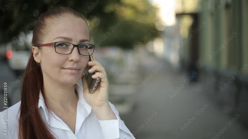 a business Caucasian middle-aged woman with red hair in optical glasses and a white blouse makes an important phone call while on the street of the city. a business Caucasian middle-aged woman with red hair in optical glasses and a white blouse makes an important phone call while on the street of the city.
