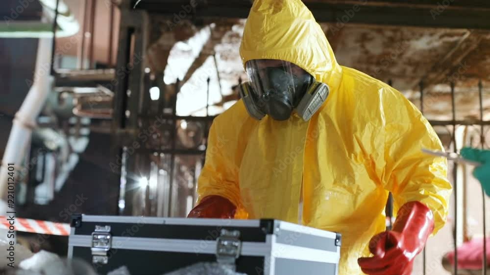 Man in gas mask and new yellow protective suit opening black briefcase. Guy wearing red rubber gloves looking for something. Indoors. Man in gas mask and new yellow protective suit opening black briefcase. Guy wearing red rubber gloves looking for something. Indoors.