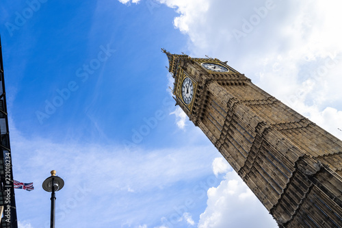 Low angle view of the Big Ben in London England United Kingdom against a blue sky with white clouds and a Union Jack British flag on a building and a streetlight
