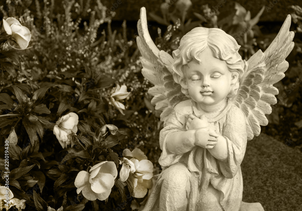 angel made of clay sitting on a grave surrounded by flowers - sepia colored