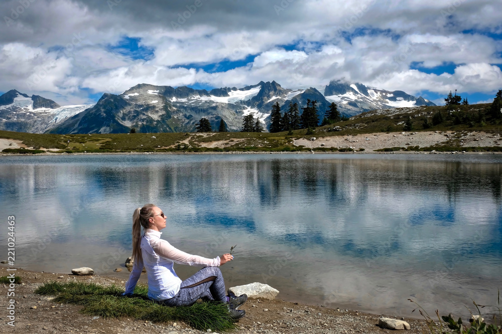 Woman on the lake shore meditating and relaxing. Beautiful view of mountains and reflections in the lake. Alpine meadows in Garibaldi Park near Whistler. British Columbia. Canada.