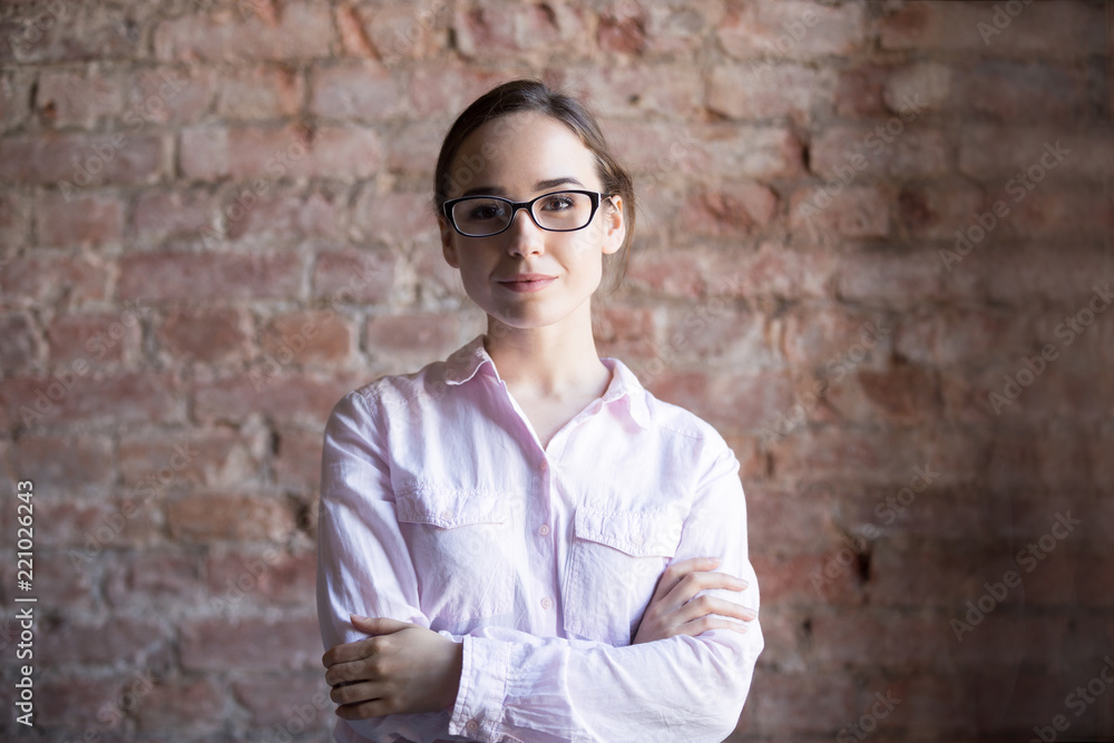 Head shot portrait of young confident professional worker woman in eyeglasses standing in office with arms crossed on chest and looking at camera